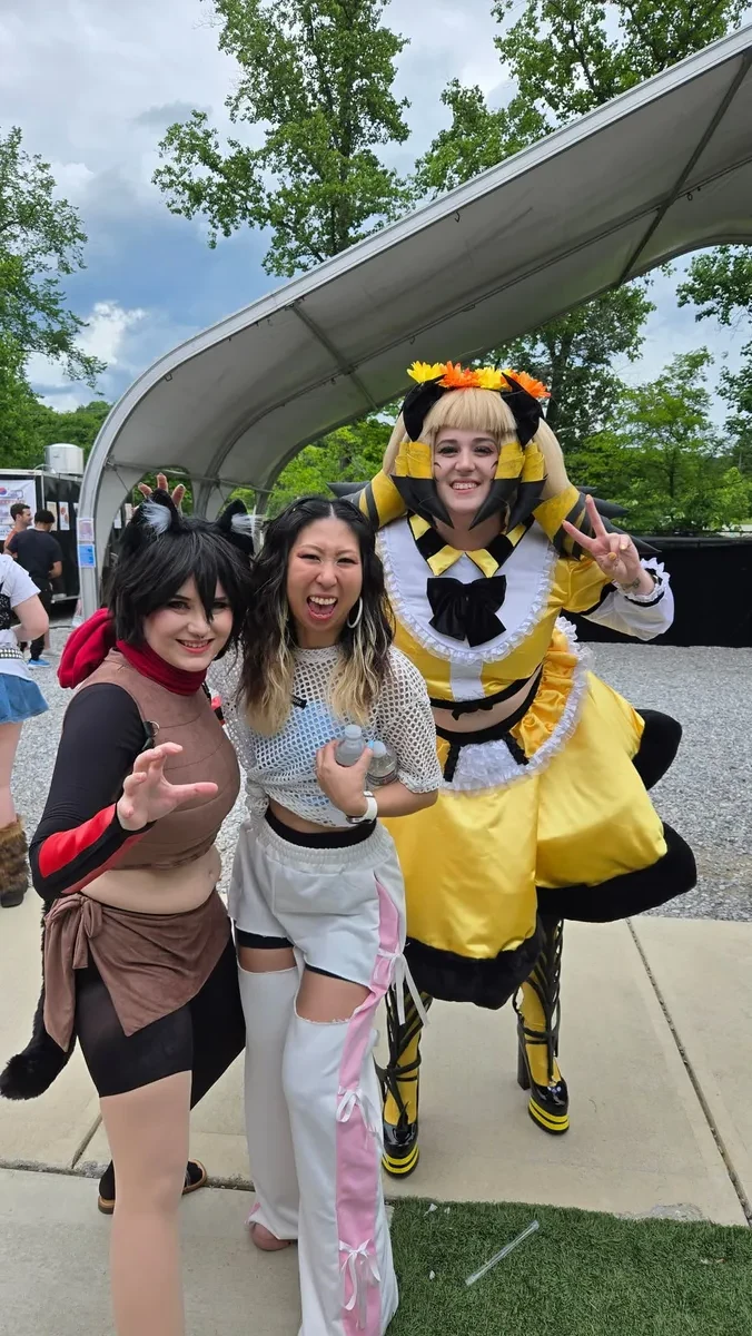 Three costumed attendees strike peace-sign poses beside a paved path and trees, one in a black-and-yellow bee-themed maid outfit, at Knox Pop Con.