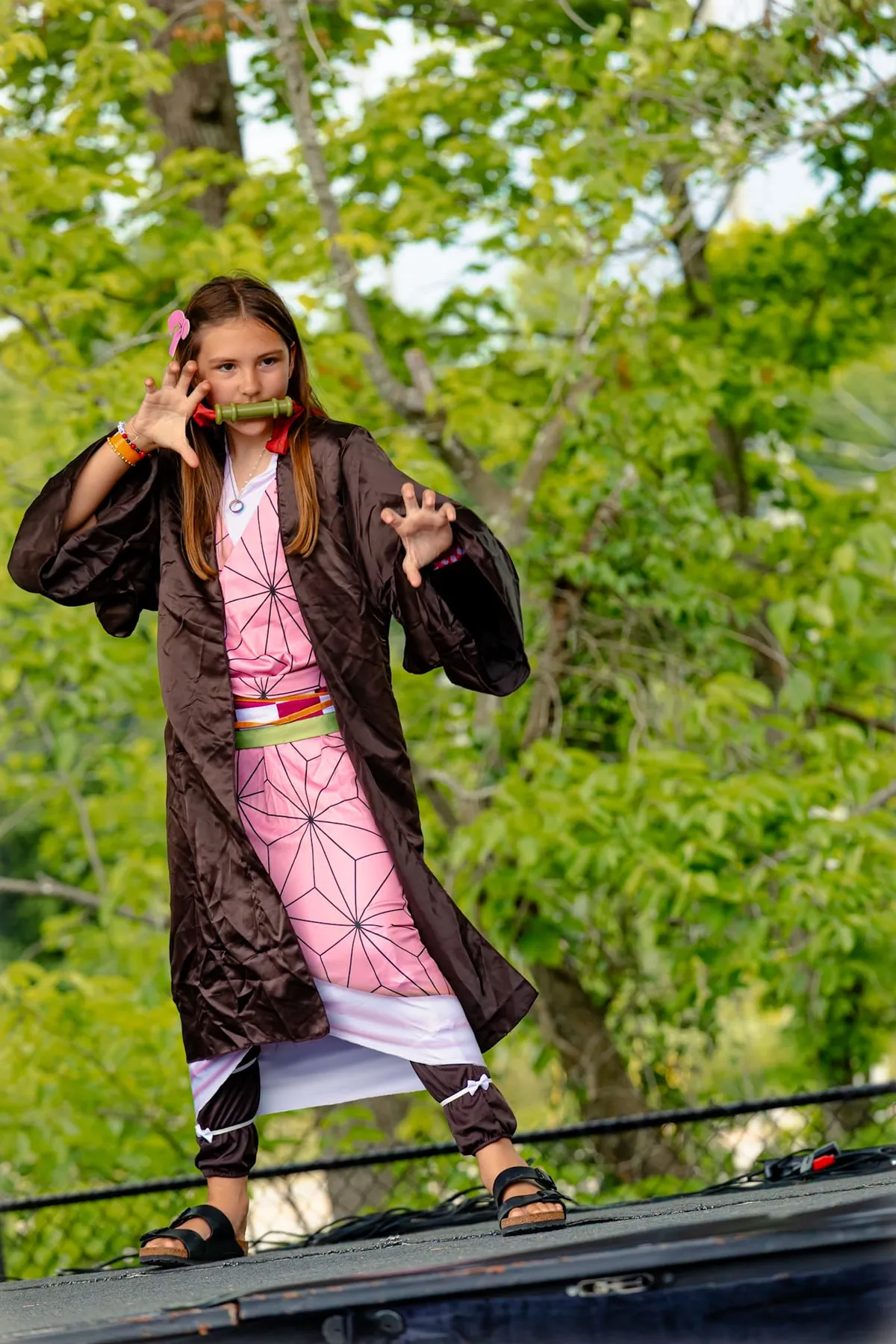 A child in a pink kimono and brown haori poses with a bamboo muzzle and clawed hands on an outdoor stage backed by green trees at Knox Pop Con.