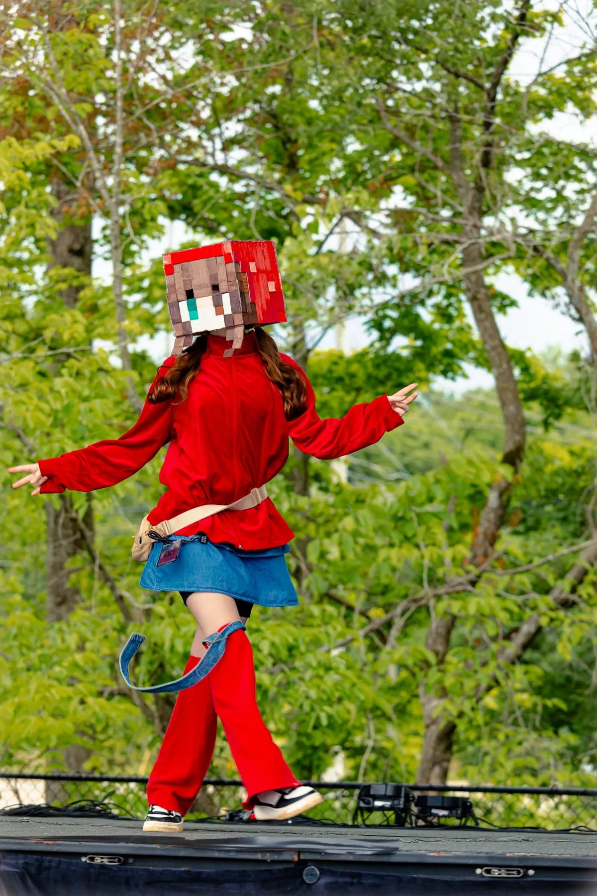 A performer wearing a blocky pixelated Minecraft-style head and red outfit dances on an outdoor stage backed by green trees at Knox Pop Con.