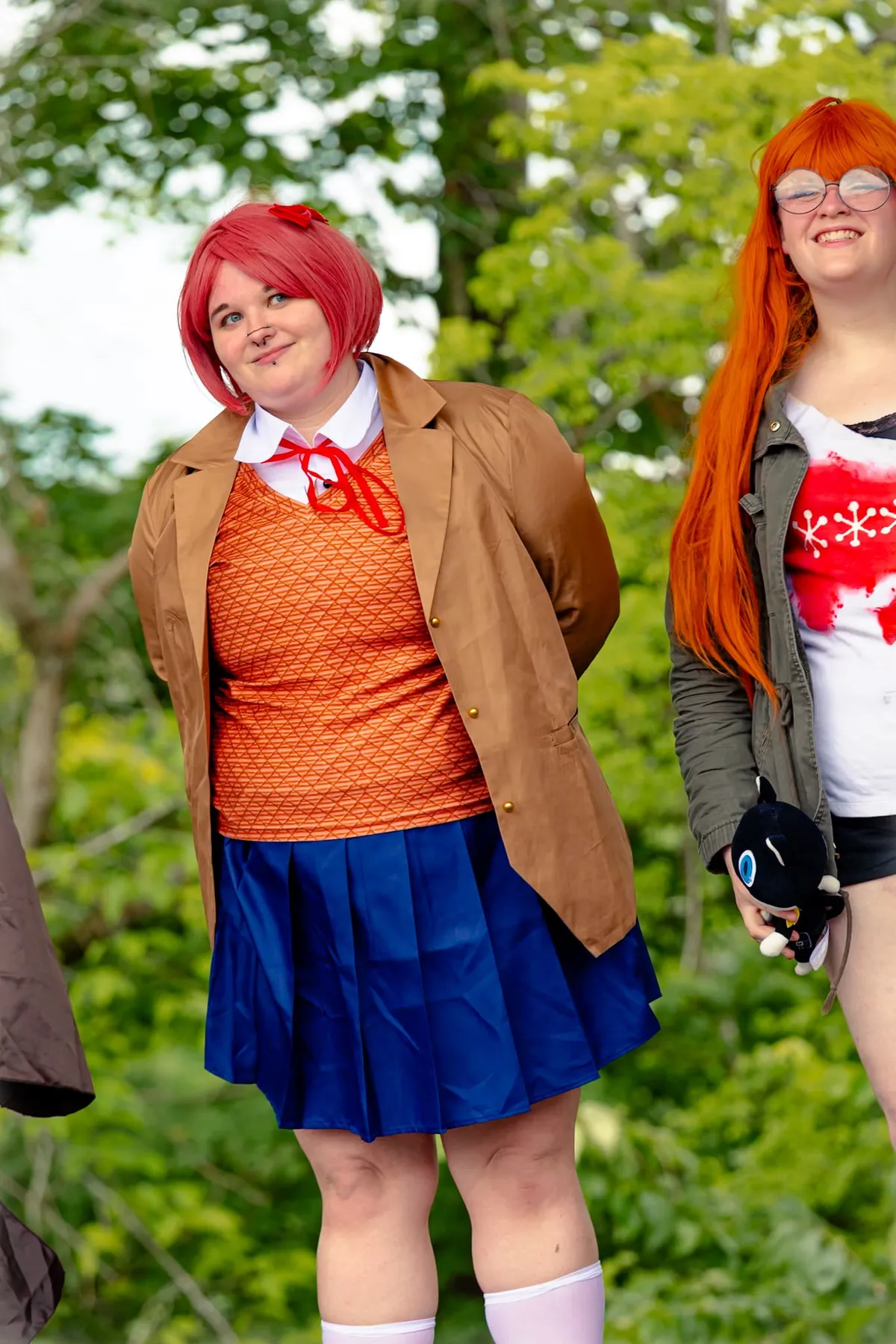 Two cosplayers pose outdoors, one with a pink bob and tan jacket and one with long orange hair holding a black-and-white cat plush, at Knox Pop Con.