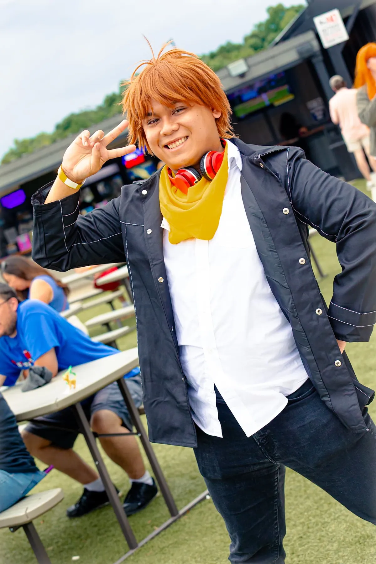 A young man in an orange wig, yellow neckerchief and red headphones throws a peace sign near picnic tables at an outdoor venue at Knox Pop Con.