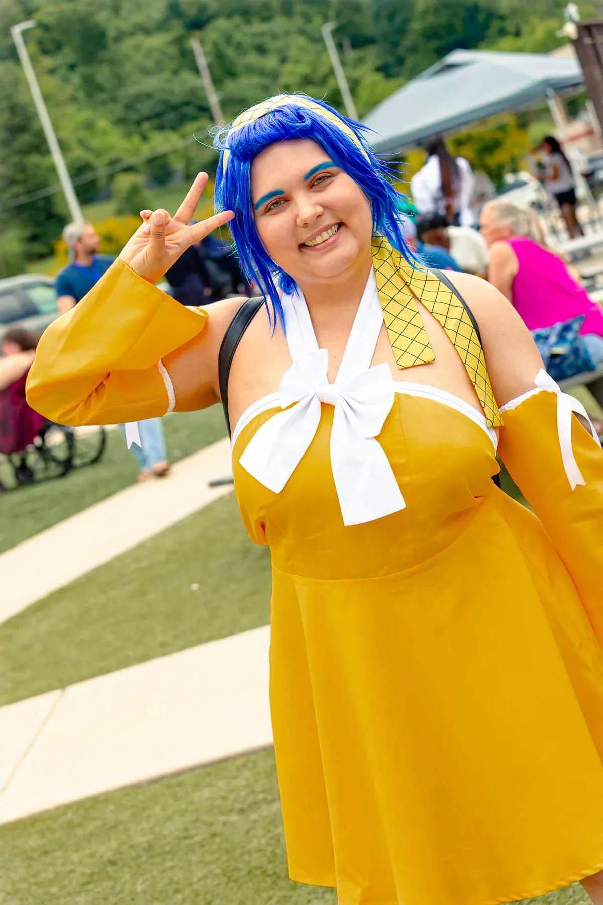A cosplayer with a bright blue wig and yellow dress with a white bow flashes a peace sign on a grassy lawn with attendees seated behind at Knox Pop Con.