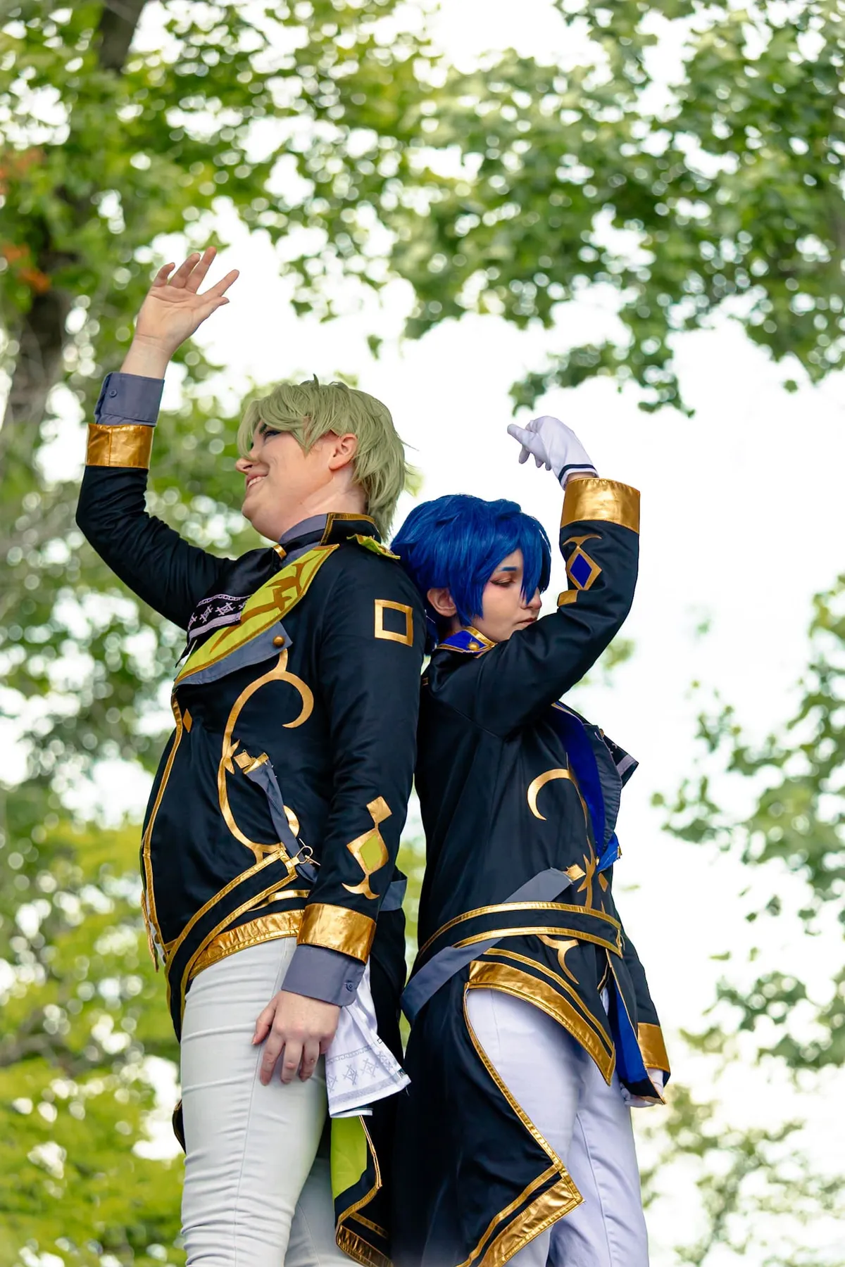 Two cosplayers in matching navy-and-gold military-style uniforms stand back to back with arms raised, one with green hair and one with blue hair, against leafy trees at Knox Pop Con.