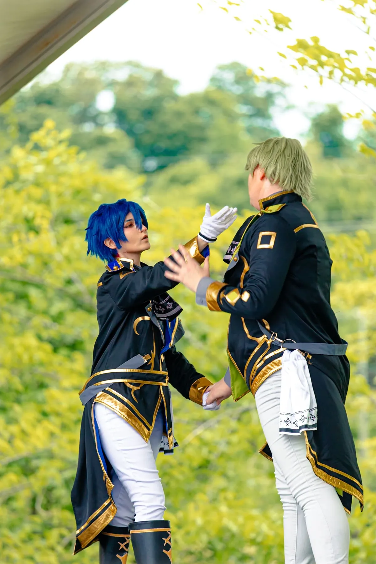Two cosplayers in navy-and-gold uniforms face each other mid-gesture, one with blue hair and one with green hair, in front of green foliage at Knox Pop Con.