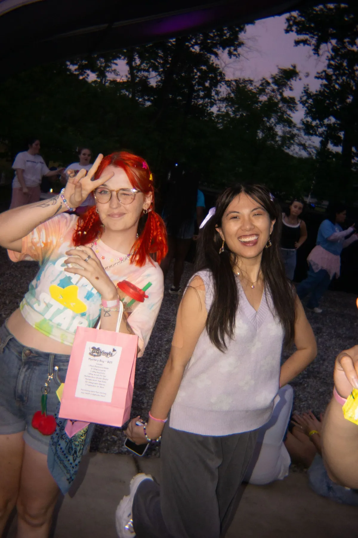Two attendees pose at dusk, one with bright red hair flashing a peace sign and holding a pink mystery-bag, the other smiling in a lavender sweater vest, at Knox Pop Con.