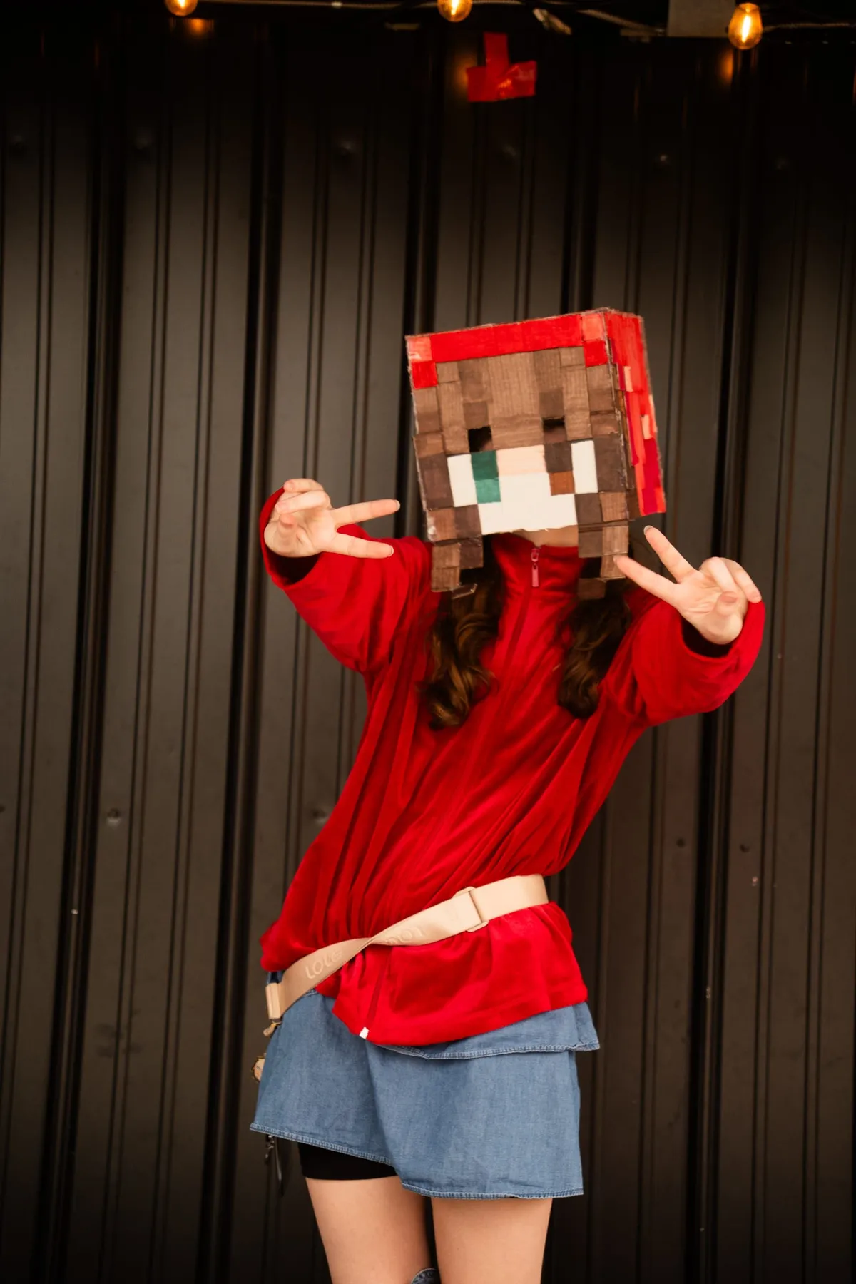 A cosplayer in a red top and denim skirt with a handmade blocky cardboard head throws double peace signs on stage at Knox Pop Con.
