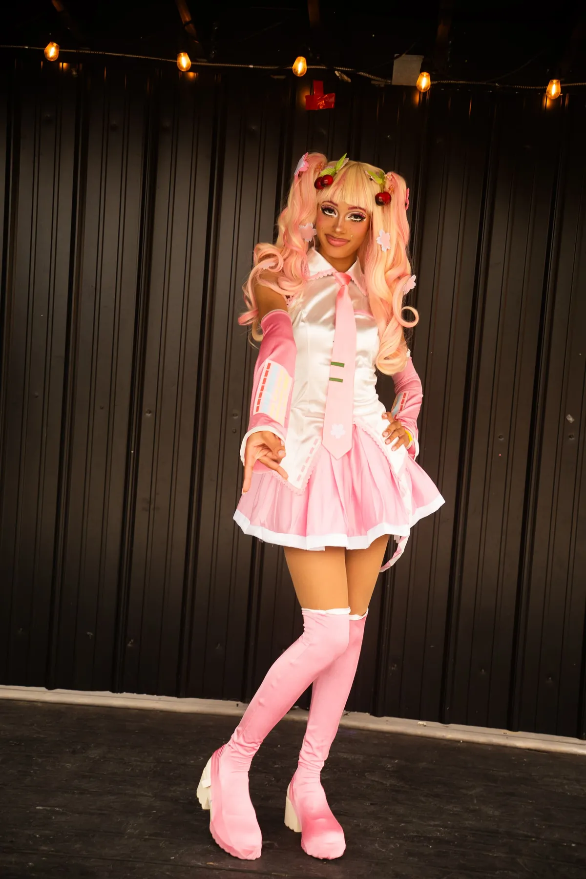 A cosplayer in a pink-and-white idol costume with long pink twin-tails, cherry hairpins and pink thigh-high boots poses full-length against a dark backdrop at Knox Pop Con.