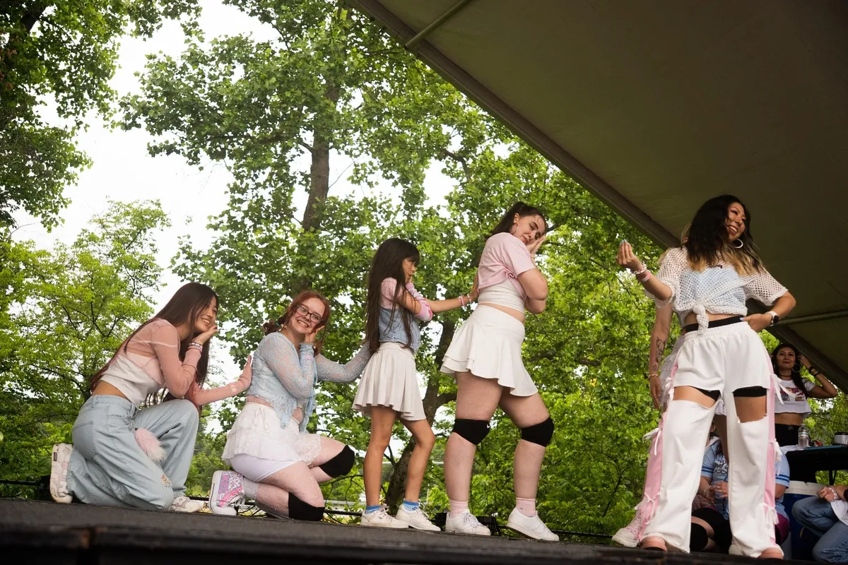 A line of performers in pastel outfits dance together on an outdoor tent stage surrounded by green trees at Knox Pop Con.