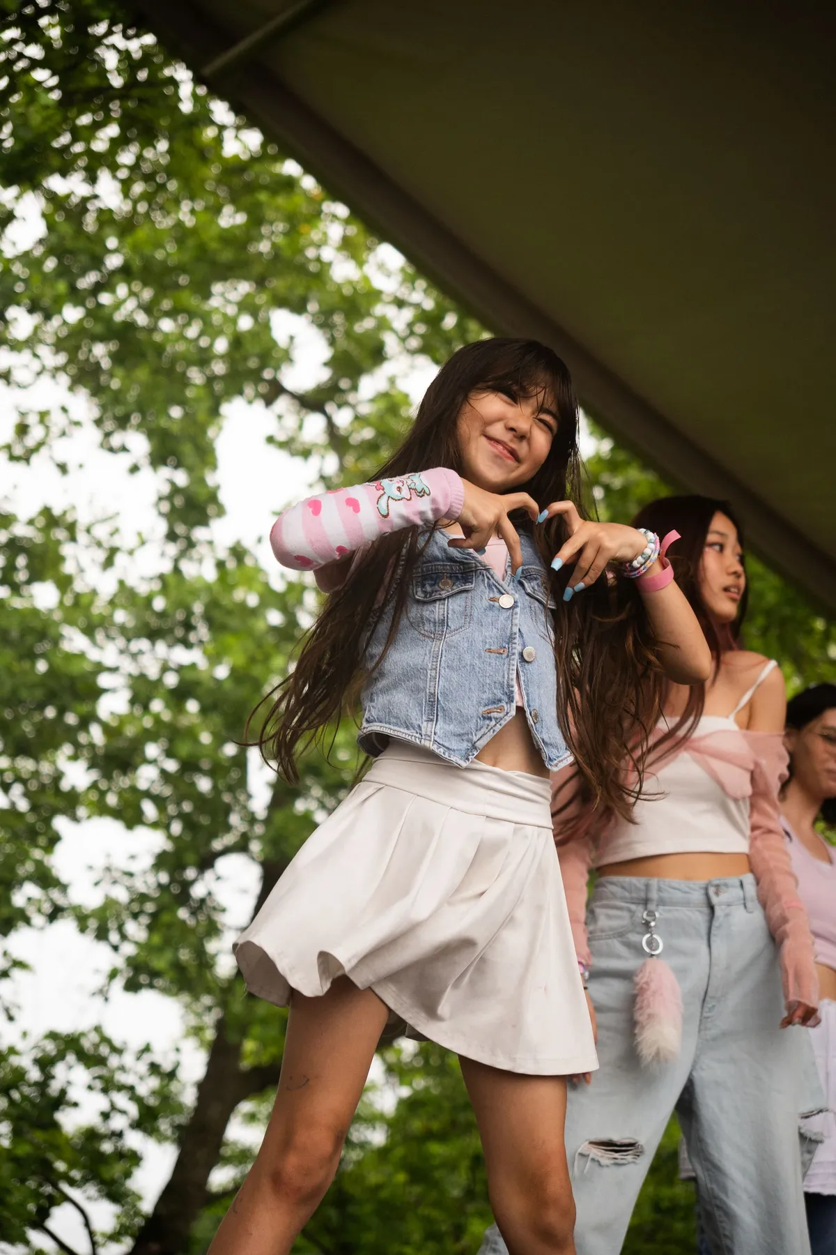 A smiling young dancer in a denim vest and white skirt forms a finger heart on an outdoor stage while another performer dances beside her at Knox Pop Con.