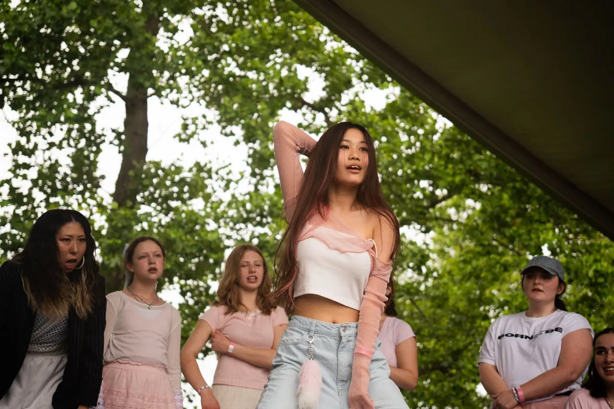 A young dancer in a white crop top and pink sleeves poses with one arm raised while a line of fellow dancers holds formation behind her on a shaded outdoor stage, at Knox Pop Con.