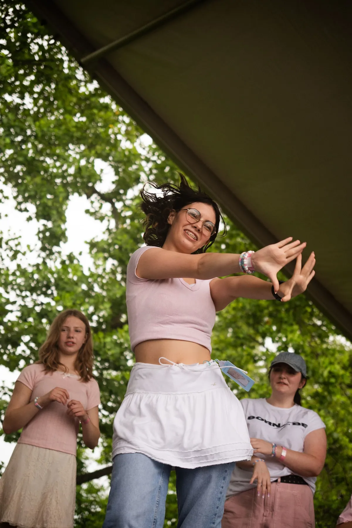 A smiling dancer in a pink top and white skirt frames a hand gesture mid-choreography while two other dancers hold position behind her on a shaded outdoor stage, at Knox Pop Con.