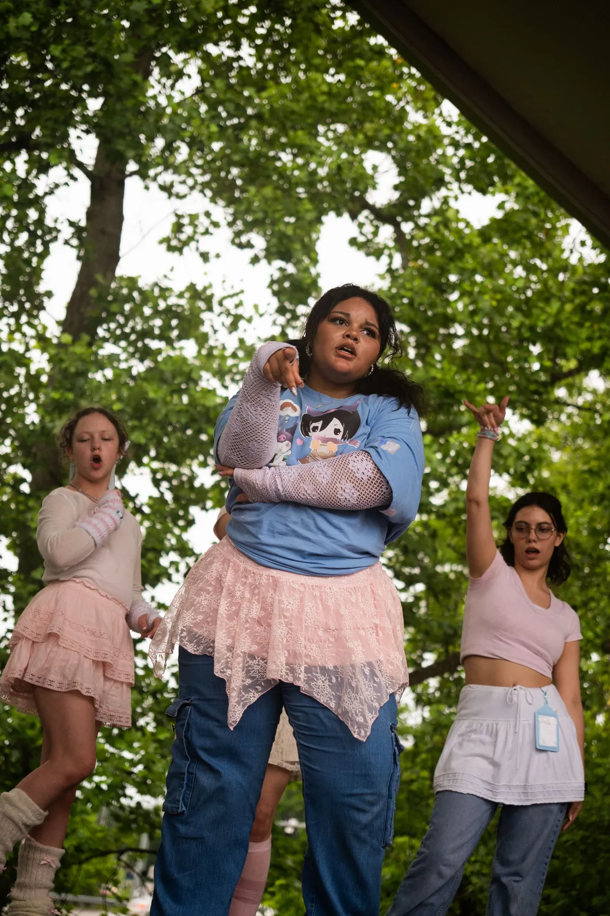 A dancer in a blue anime graphic tee and pink lace skirt points forward mid-routine while two dancers strike poses on either side on a shaded outdoor stage, at Knox Pop Con.
