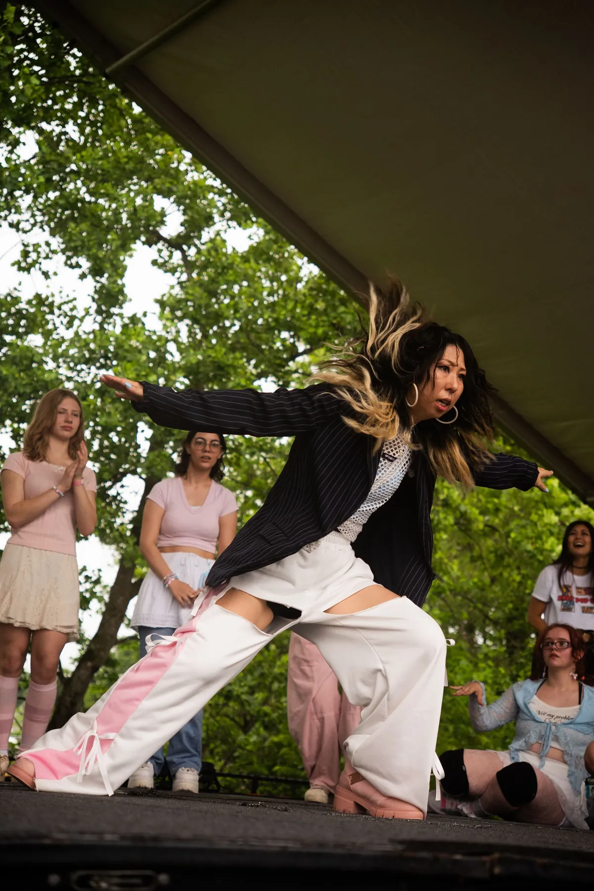 A dancer in a pinstripe blazer and white pants lunges into a dynamic move with her hair mid-flip as fellow performers watch and pose around her on a shaded outdoor stage, at Knox Pop Con.