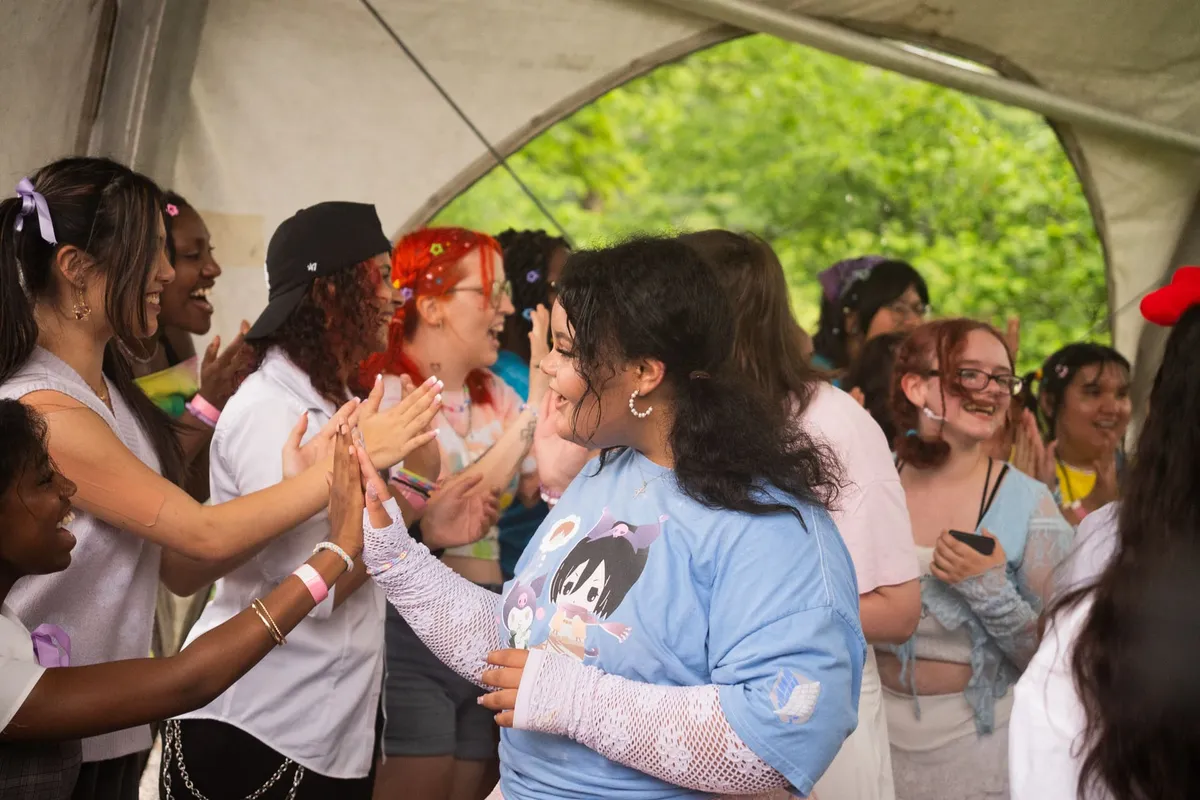 A crowd of attendees laughs and exchanges a high-five under a large tent, with a person in a light-blue anime graphic tee grinning at the center, at Knox Pop Con.