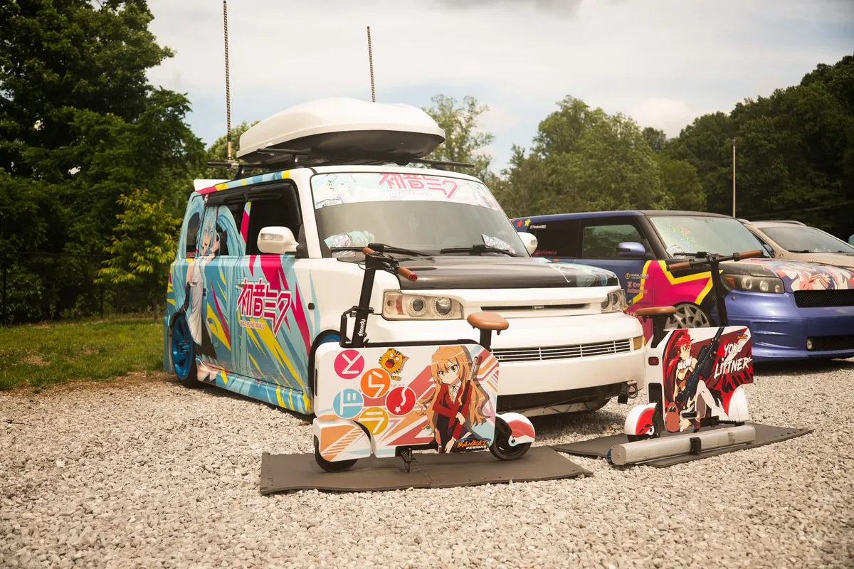 A boxy car wrapped in a teal Hatsune Miku anime design sits on a gravel lot with two anime-decorated scooters parked in front and more wrapped cars lined up alongside, at Knox Pop Con.